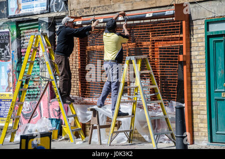 A couple of shopfitters attach a shutter to the outside of a shopfront in East London. Stock Photo