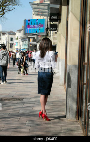 Back view of attractive slender woman with long brunette hair standing outside in a street texting in red high heels and denim skirt UK  KATHY DEWITT Stock Photo