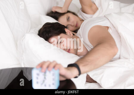 Sleepy angry couple in bed turning off the alarm clock Stock Photo