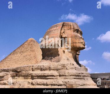 Profile view of the Great Sphinx of Giza with one of the Pyramids behind and a blue sky and wispy clouds in background in dynamic high contrast color Stock Photo