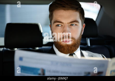 Thoughtful business man sitting in back seat of a car with newspaper in hand while looking away Stock Photo