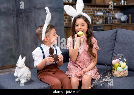 Little boy and girl in bunny ears sitting on sofa and eating sweets Stock Photo