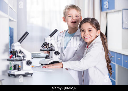 Little boy and girl in white coats using microscope and smiling at camera in lab Stock Photo