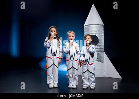 Three cute kids in astronaut costumes eating sweets standing near rocket Stock Photo