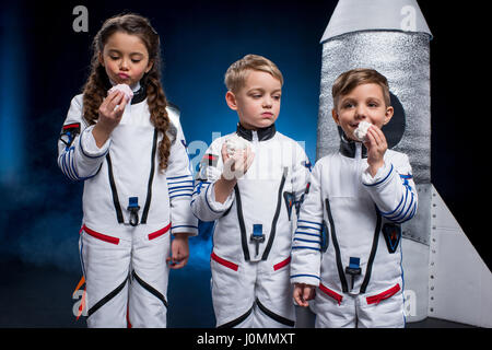 Three cute kids in astronaut costumes eating sweets standing near rocket Stock Photo