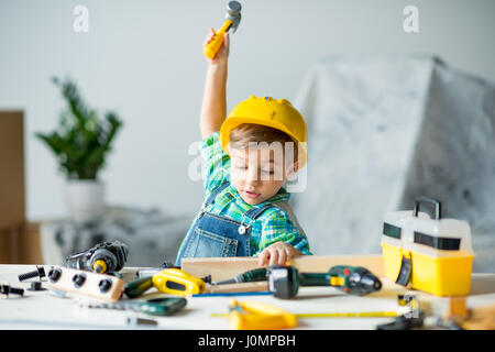 Cute little boy in yellow hard hat holding wooden plank and toy hammer Stock Photo