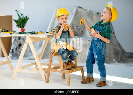 Two little boys in hard hats eating sandwiches and chatting in workshop Stock Photo