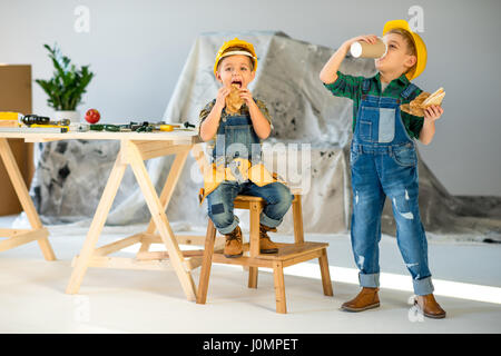 Two little boys in hard hats and denim overalls eating 