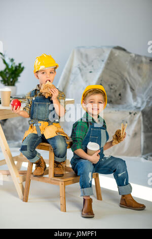 Two little boys in hard hats and denim overalls eating sandwiches in workshop Stock Photo