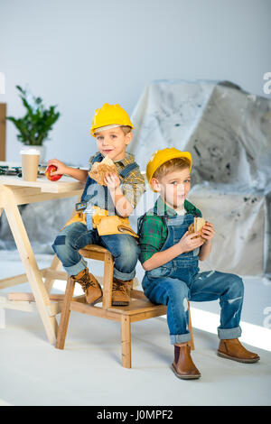 Two little boys in hard hats and denim overalls eating sandwiches in workshop Stock Photo