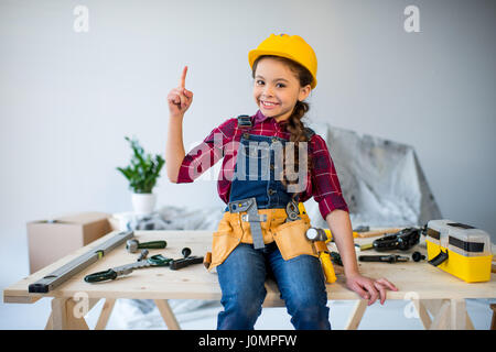 Little girl in tool belt and hard hat pointing up and smiling at camera in workshop Stock Photo