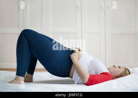 Young woman lying on bed trying to button jeans. Stock Photo