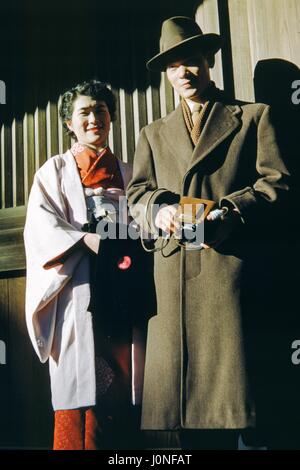 Two Western visitors to Japan pose for a photograph, the man wearing a trench coat and hat and holding a camera, the woman wearing a Japanese kimono, both with partial smiles and standing in shadow, Japan, 1952. Stock Photo