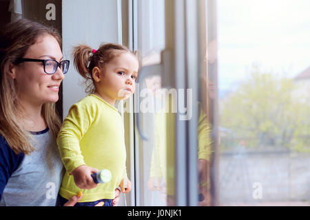 A happy family. Little girl looks out the window with mother Stock Photo