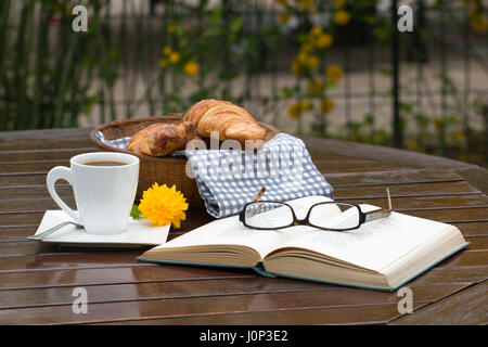 A basket with croissants, a cup of coffee, an open book and a pair of eyeglasses on a wooden table Stock Photo