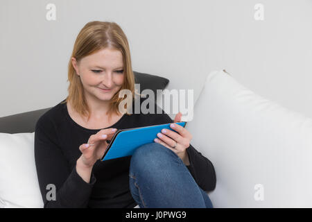 Closeup view of smiling young woman using a tablet sitting on the black and white couch with black and white pillows. Potential trademarks and buttons Stock Photo