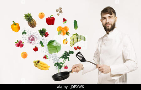 Handsome professional chef in uniform uggling with vegetables and other food in the kitchen. Chef and flying vegetables and fruits osilated on white b Stock Photo