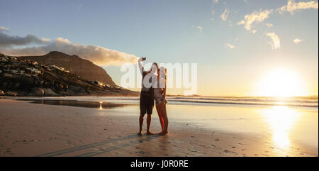 Horizontal shot of loving young couple taking self portrait at the beach. Man and woman on sea shore taking selfie with mobile phone. Stock Photo