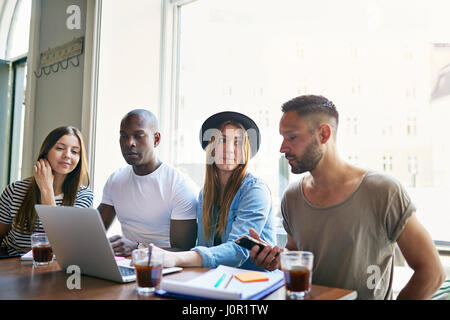 Group of four young co-workers sitting at table with laptop and having coffee while discussion in office. Stock Photo