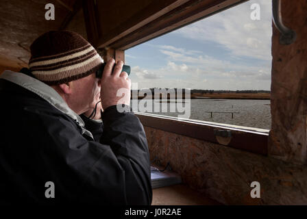 A man sitting in a viewing hide looking through binoculars at birds Stock Photo