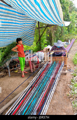 Woman working at loom, weaving extended rug  (18 meters) to be section into smaller lengths, young daughter observing, along rural road. Stock Photo