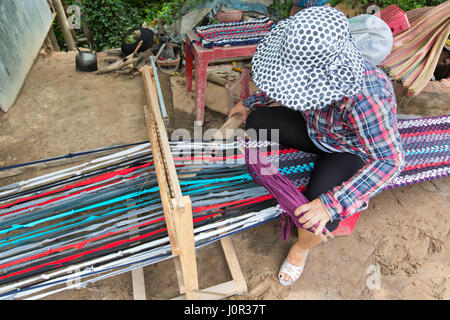 Woman working at loom,  holding spindle, weaving extended rug  (18 meters) to be section into smaller lengths, along rural road. Stock Photo