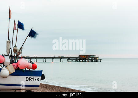 England, Deal beach. Stern of fishing boat on shingle, and in back ground, deal concrete Pier. Sea is calm, dawn sky, faint band of pink of horizon, grey could above that. Stock Photo