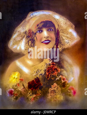 Hand-colored vintage portrait of a smiling 1920s era Flapper girl in her twenties wearing a wide brim cloche hat holding a bouquet of carnation flower Stock Photo