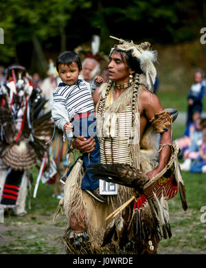 A Native American Indian father dressed in tribal costume dancing at a powwow holding his young son. Hanover, NH, USA. Stock Photo