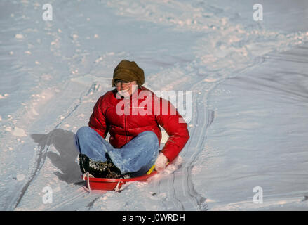 Adult woman in a bright red jacket slides down a hill on a plastic snow sled in Plainfield, New Hampshire, United States. Stock Photo