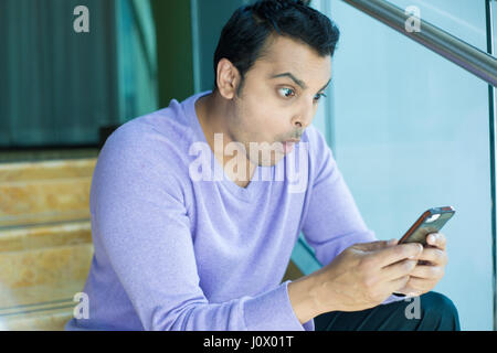 Closeup portrait, funny young man, shocked surprised, wide open mouth, large eyes by what he sees on his cell phone, isolated indoors office backgroun Stock Photo