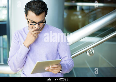 Closeup portrait, young captivated, absorbed, engrossed man in purple sweater and black eye glasses perusing, pondering emails on silver gray tablet t Stock Photo