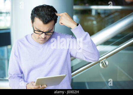 Closeup portrait, dumbfounded, flabbergasted man in black eyeglasses and purple sweater, scratching head, looking at tablet, isolated indoors office b Stock Photo
