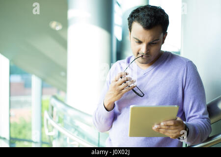 Closeup portrait, young captivated, absorbed, engrossed man in purple sweater biting black eye glasses, perusing, pondering emails on silver gray tabl Stock Photo