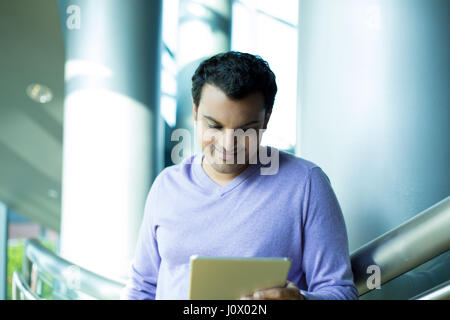 Closeup portrait, young captivated, absorbed, smiling man in purple sweater perusing, pondering emails on silver gray tablet touch-pad, isolated indoo Stock Photo