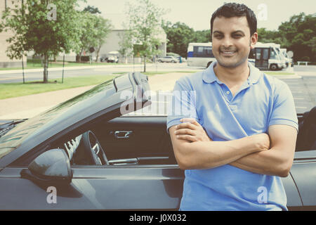Closeup portrait, young handsome man in blue polo standing in front of his gray charcoal black sports car, arms crossed folded, isolated on outdoors b Stock Photo