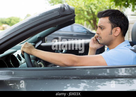 Closeup portrait, sad handsome young man in blue polo shirt sitting, driving in gray black sports car on cellphone with bad news, isolated outdoors ba Stock Photo