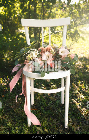 Bridal bouquet of pink flowers and greenery with ribbon, lies on vintage white wooden chair Stock Photo