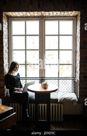 silhouette of young woman sitting at table in front of window and uses laptop. Stock Photo