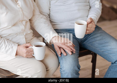 Always together. Close up of a nice loyal elderly couple sitting together and holding hands while having tea Stock Photo
