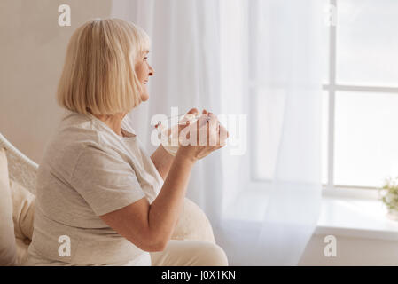 Tasty meal. Pleasant positive senior woman having breakfast and smiling while looking into the window Stock Photo