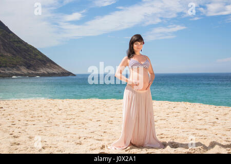 Pregnant woman sitting on beach cradling her belly Stock Photo