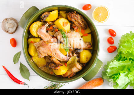 Top flat view of roasted rabbit meat with vegetables and herbs in round ceramic pot on white wooden table surface Stock Photo