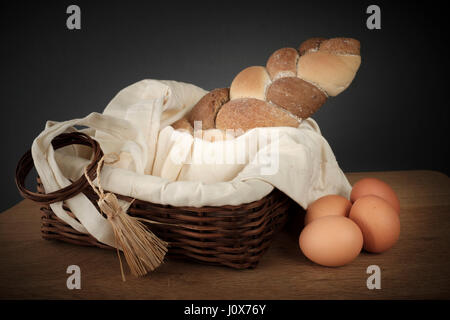 3 types interwoven bread in a wicker basket beside a few eggs on a wooden table Stock Photo