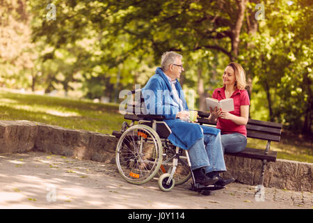 Young carer woman reading book outdoor disabled senior man in wheelchair Stock Photo