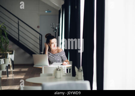 Beautiful young woman making notes in her laptop and looking through window while sitting at a table in a cafe. Stock Photo