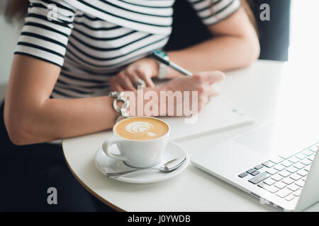 Close up picture of a cup of cappuccino with a pattern and hands writing in a notebook. Woman sitting in a cafe woth a laptop and making notes. Stock Photo