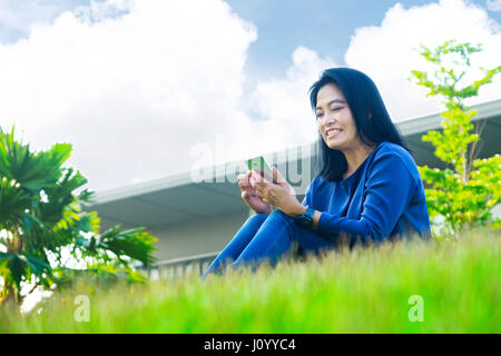 Asian woman chatting on mobile phone and smiling at screen sitting on grass outside home,Low angle view. Stock Photo