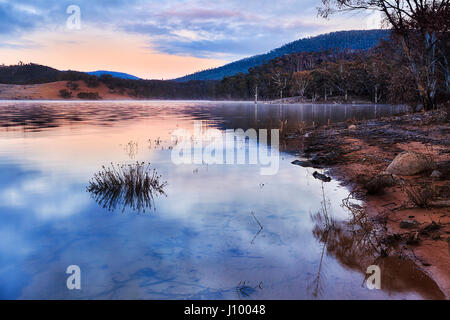 Pink sunrise over hills change around Jindabyne lake in Snowy mountains national park of Australia. Rising sun reflects in still water of winter lake. Stock Photo