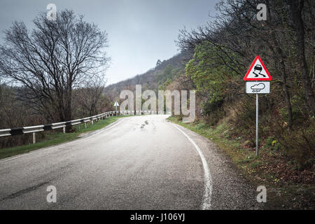 Slippery road, warning roadsign stands on mountain roadside, rainy rural landscape Stock Photo